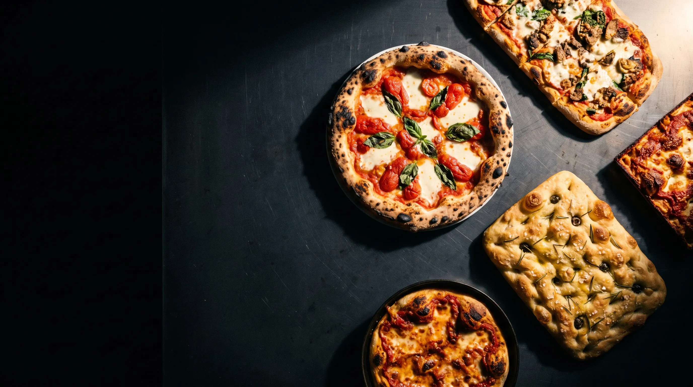 Five styles of pizza arranged on a dark steel bench.